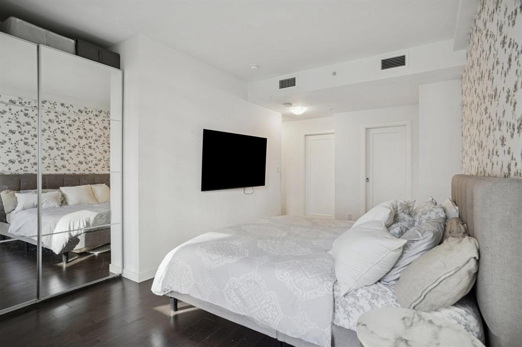 Bedroom featuring a large bed with white bedding, patterned wallpaper, mirrored closet, wall-mounted TV, and dark wood floors.