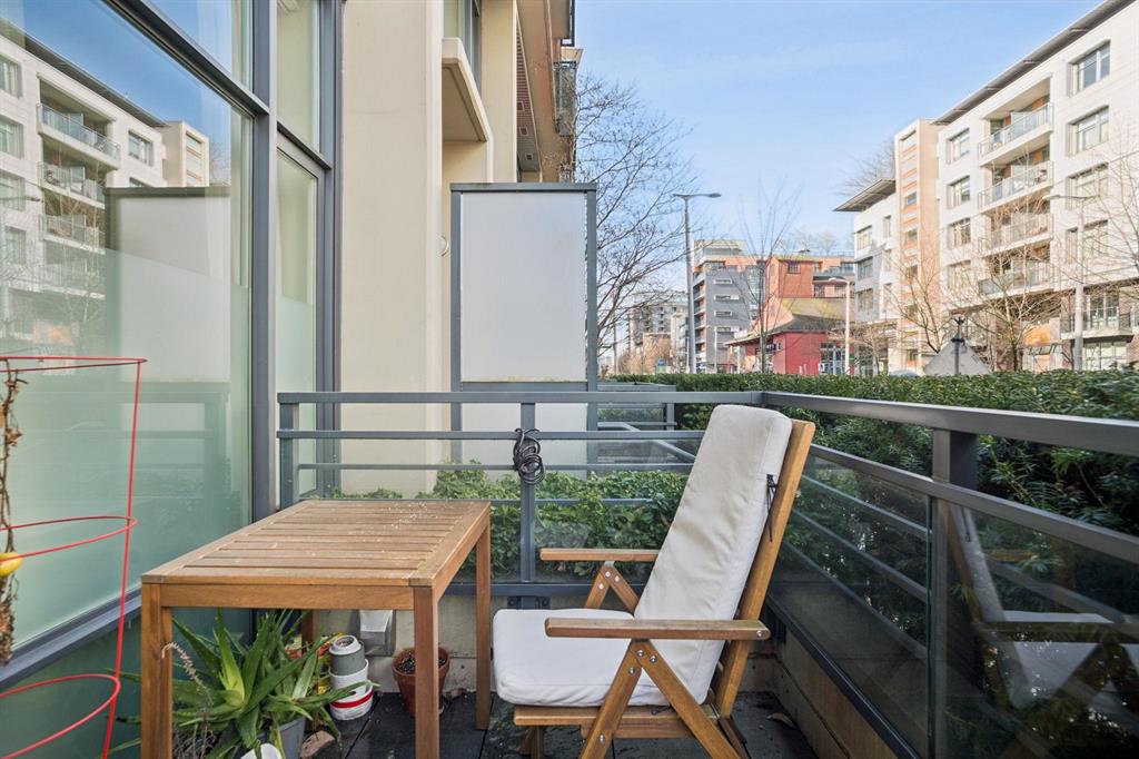 A cozy balcony with a wooden table, cushioned chair, and potted plants overlooking modern apartments across the street.