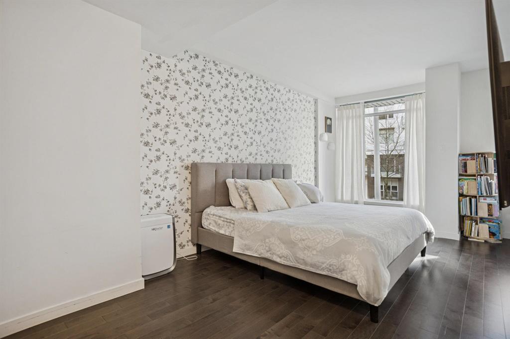 Modern bedroom featuring a gray upholstered bed, floral accent wall, white bedding, air purifier, light curtains, and corner bookshelf.