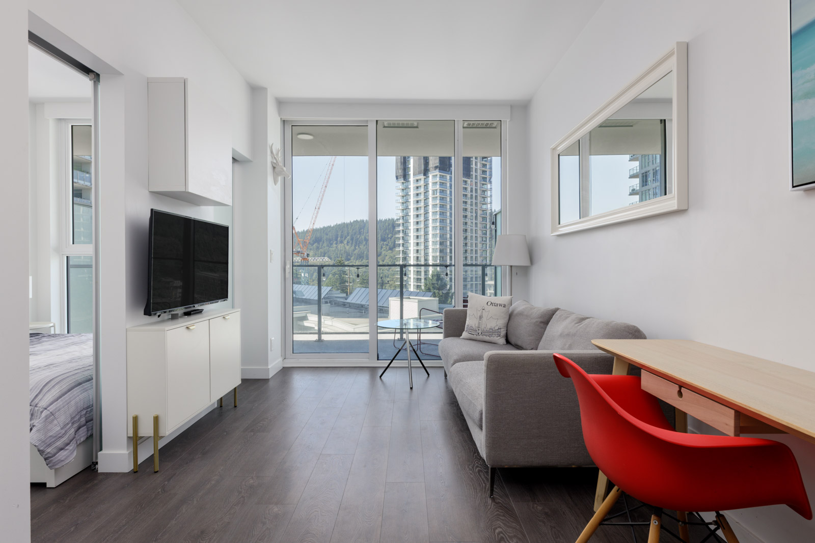 Living room at 567 Clarke Rd, Coquitlam with gray sofa, TV, wooden desk and red chair, large window, balcony view; bedroom to left.