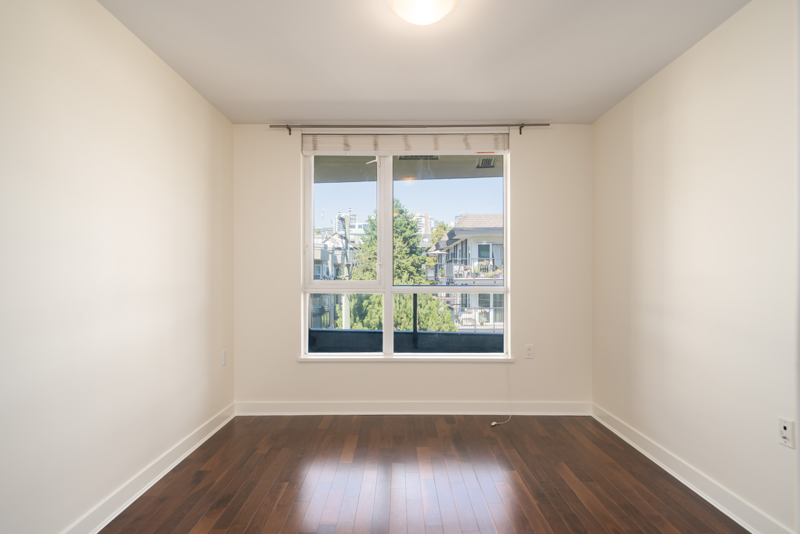 Empty room with pale walls, dark wood floors, large window facing trees and buildings. 5th floor, 160 3rd St W, North Vancouver.