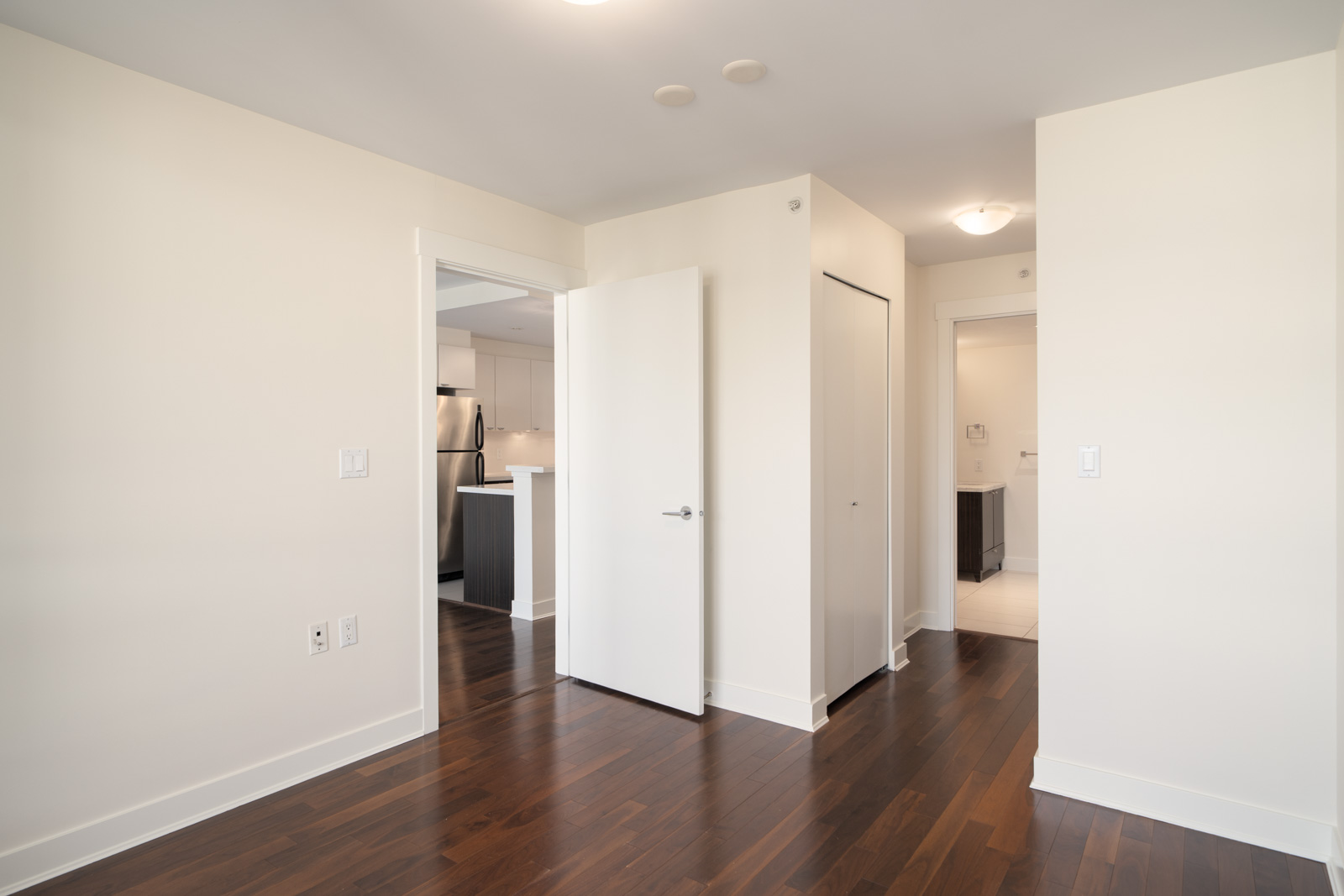 Bright empty room with dark wood floors, white walls, open door to kitchen, closed closet, and hallway and bathroom in background. North Vancouver.
