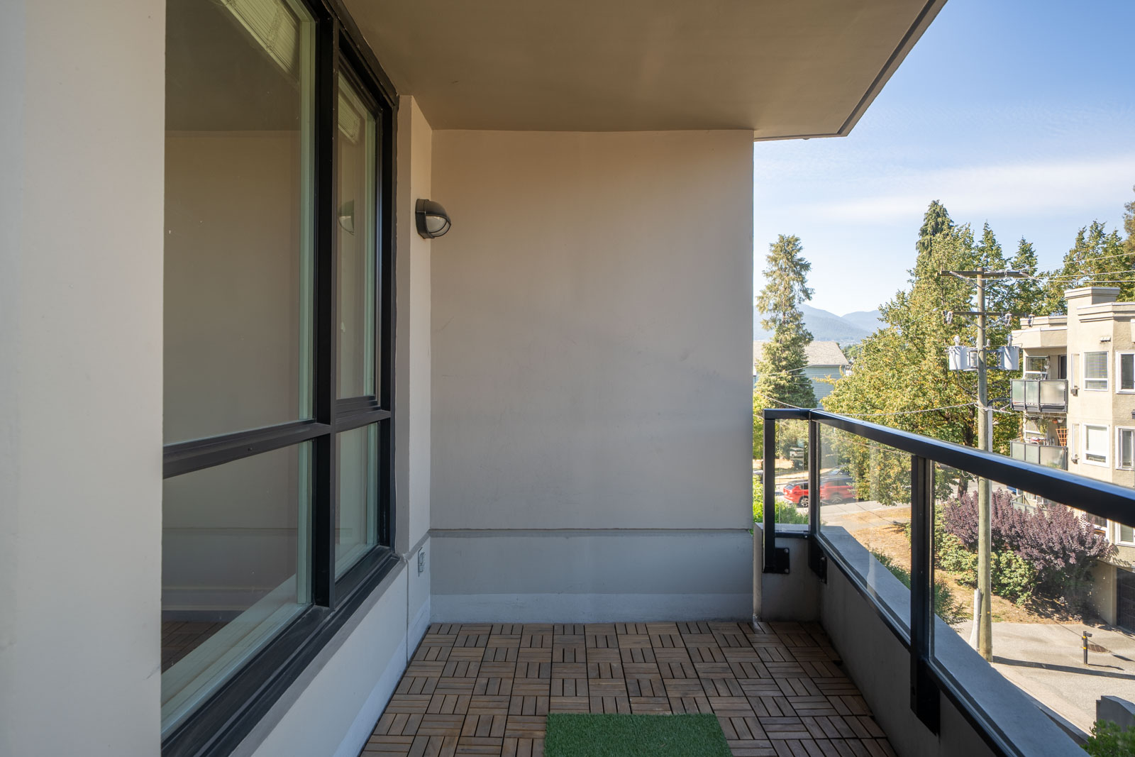 Apartment balcony on 5th floor with wood tile floor, glass railing, and views of trees, buildings, and blue sky in North Vancouver.