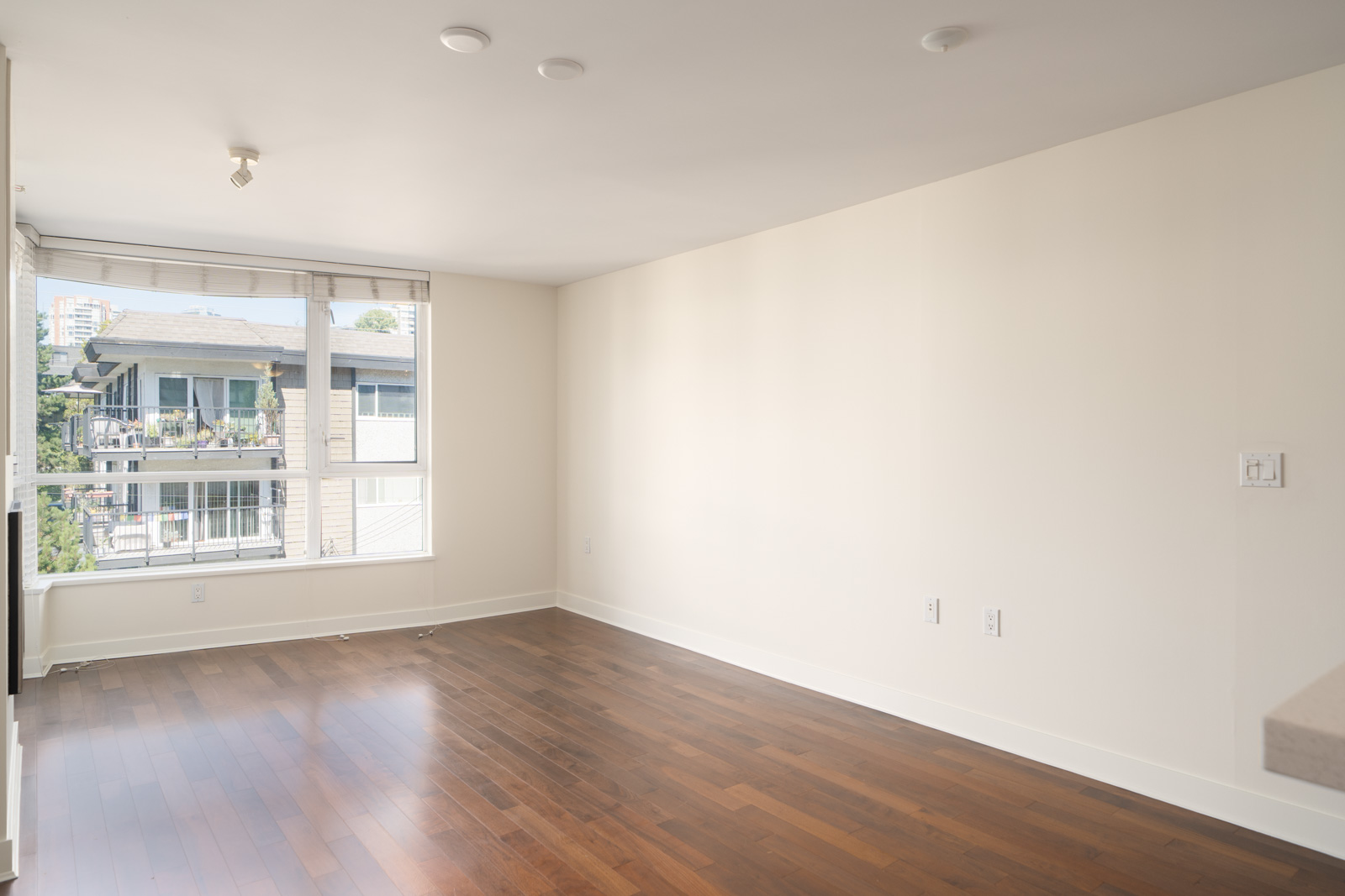 Empty room with white walls, hardwood floor, large window, and sunlight overlooking North Vancouver buildings from the 5th floor.