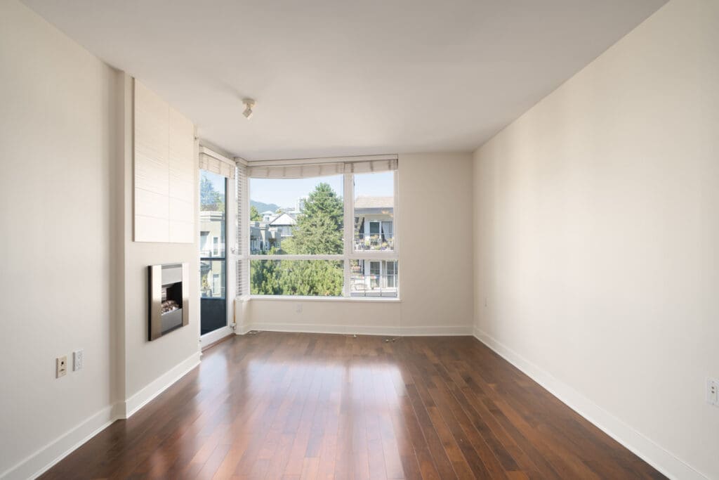 Empty room with hardwood floors, large windows, light walls, and a small fireplace on 3rd St W, North Vancouver, fifth floor.