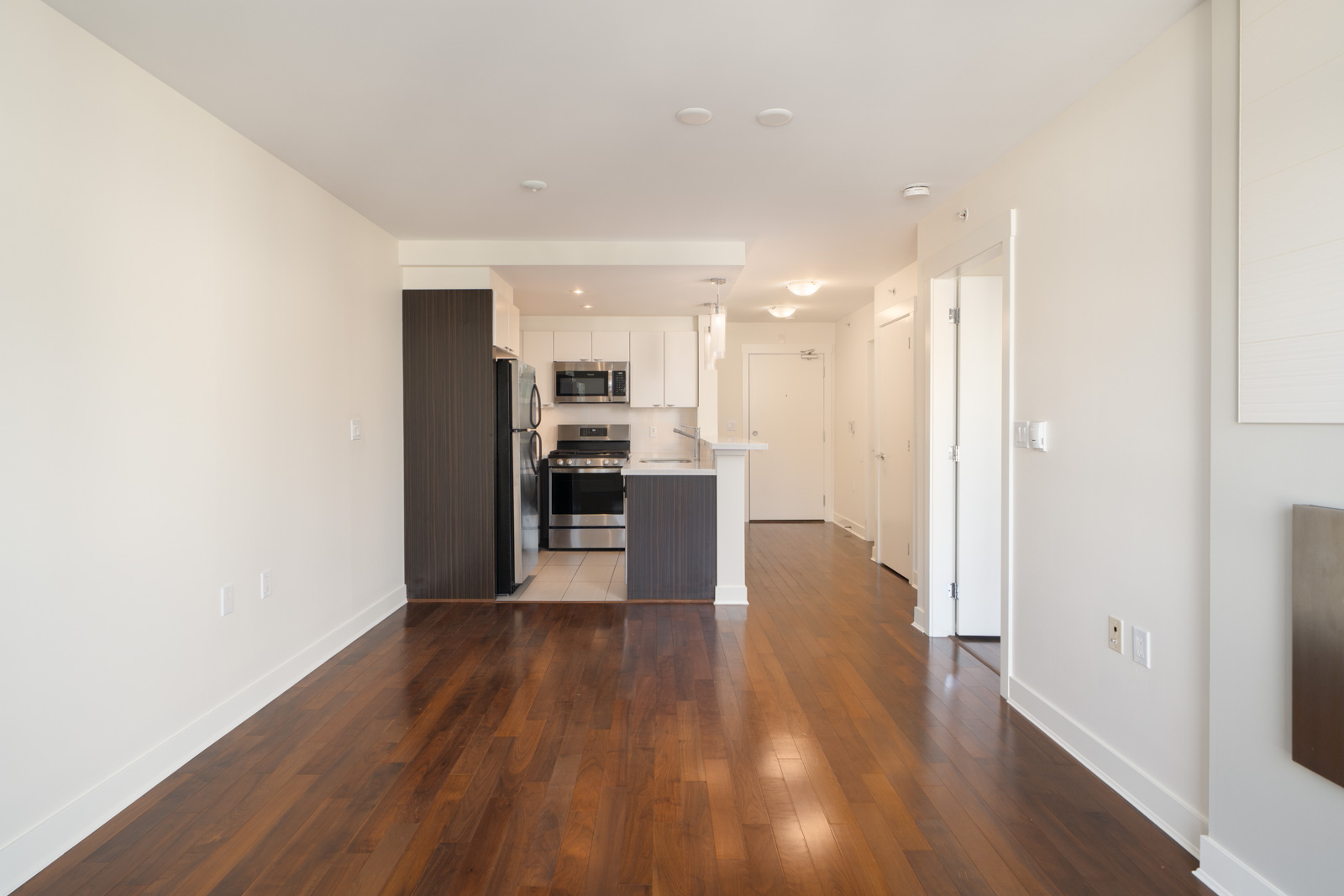 Empty apartment at 160 3rd St W, North Vancouver with hardwood floors, white walls, modern dark kitchen, and visible hallway.