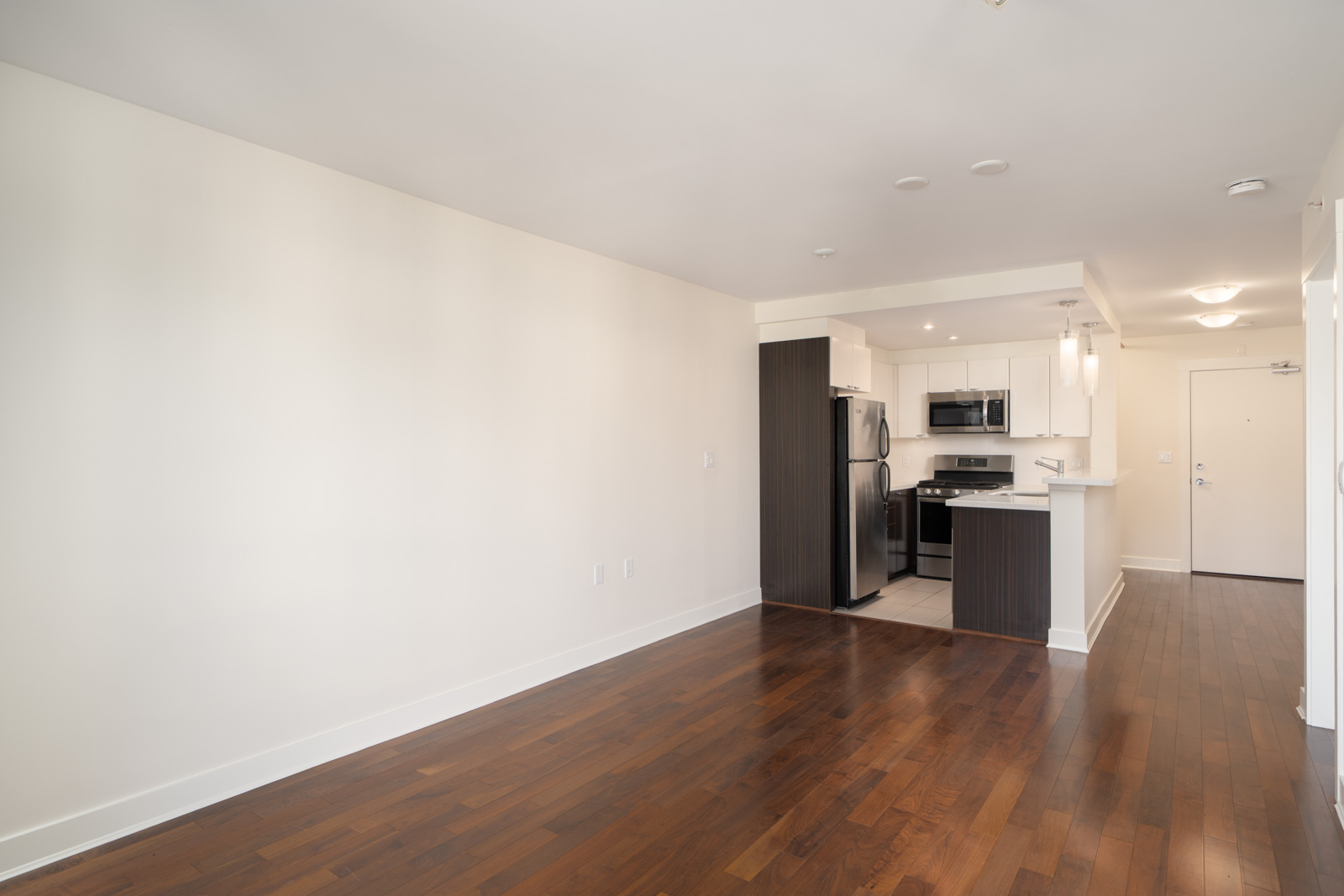 Empty apartment with hardwood floors, white walls, open kitchen with stainless appliances, and entry door. North Vancouver.