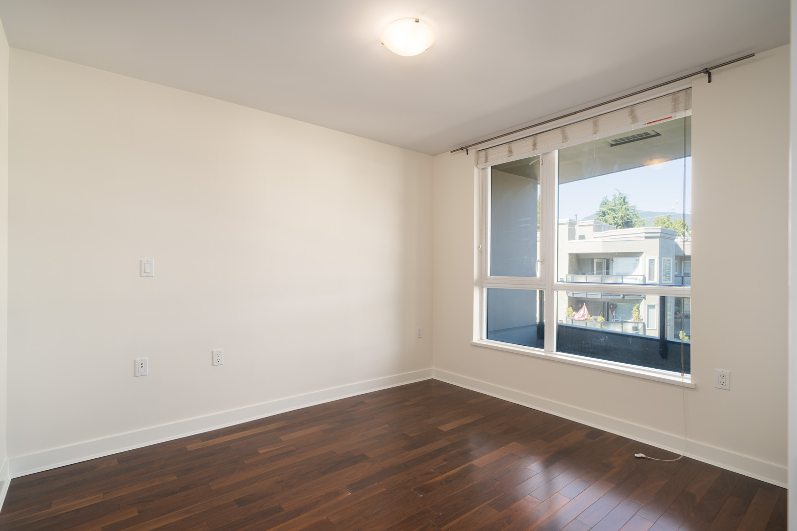 Empty room with white walls, hardwood floor, ceiling light, and large window facing balcony and buildings—5th Floor, North Vancouver.