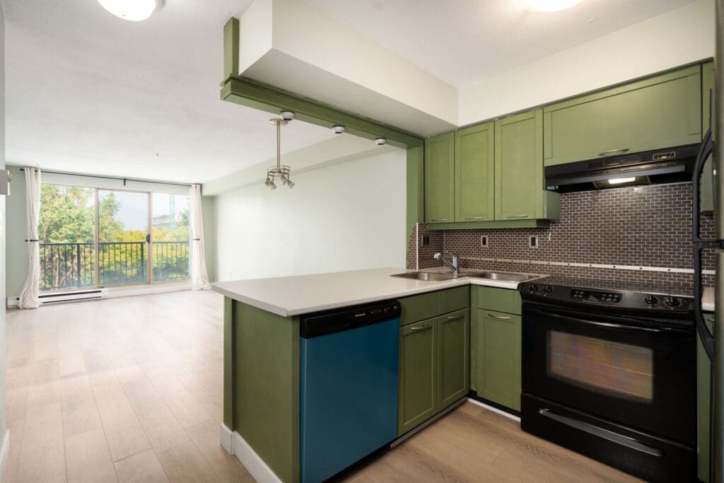 Modern kitchen with green cabinets, black appliances, blue dishwasher; opens to bright living area and balcony at 1099 E Broadway, Vancouver.