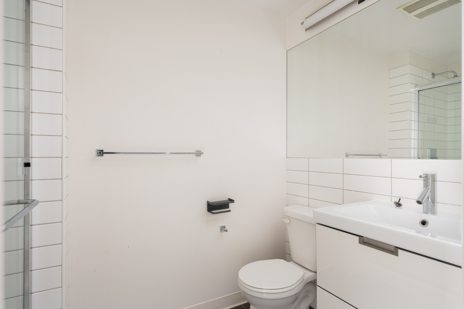 Minimalist bathroom at 138 E Hastings St, Vancouver with white walls, toilet, sink, large mirror, glass shower, and wall-mounted fixtures.