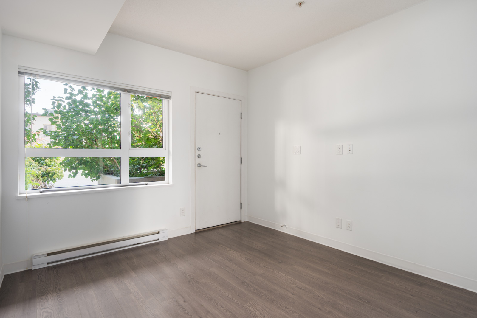 Empty room with white walls, dark wood floor, large window showing trees, and closed door at 138 E Hastings St, Vancouver.