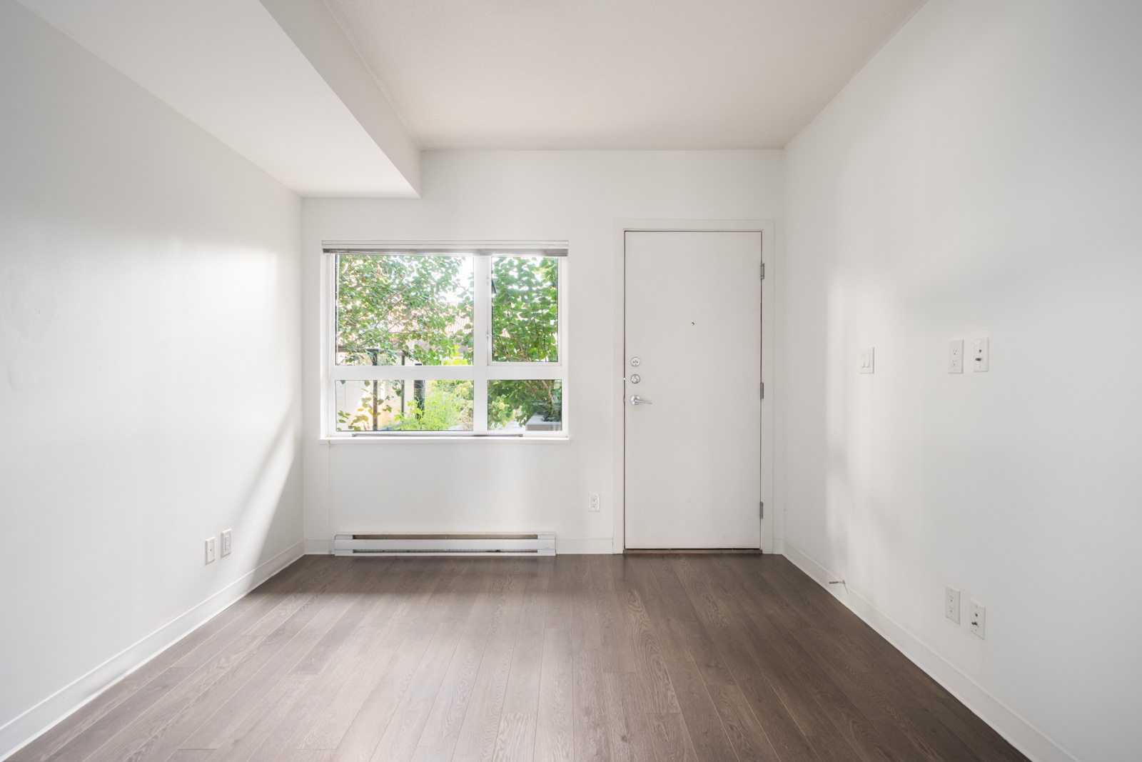 Empty room with white walls, dark wood floor, window with greenery view, white door, and few outlets on right wall in Vancouver.