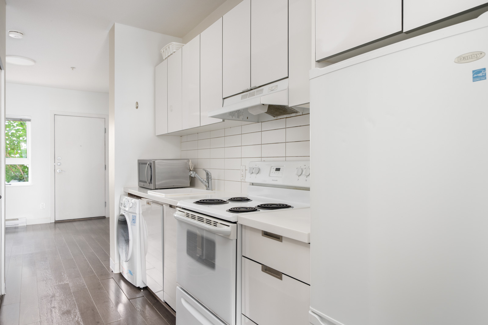 Narrow kitchen with white cabinets, electric stove, fridge, microwave, washer; dark wood floor and tiled backsplash in Vancouver.
