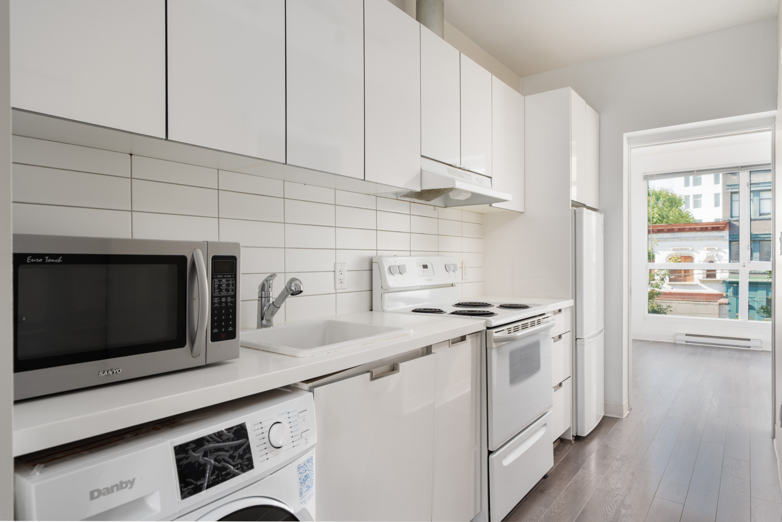 Modern kitchen with white cabinets, electric stove, microwave, washer, fridge, and tile backsplash; sunlight from adjacent room in Vancouver at 138 E Hastings St.