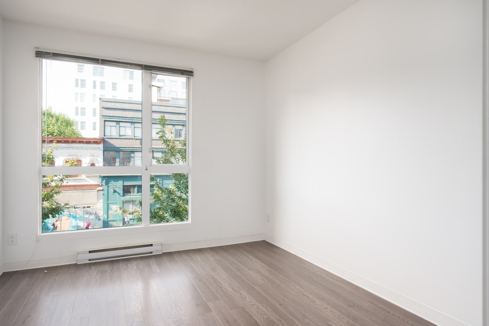 Empty room with white walls, large window, and dark wood floor on 2nd floor; view of buildings and trees in Vancouver.