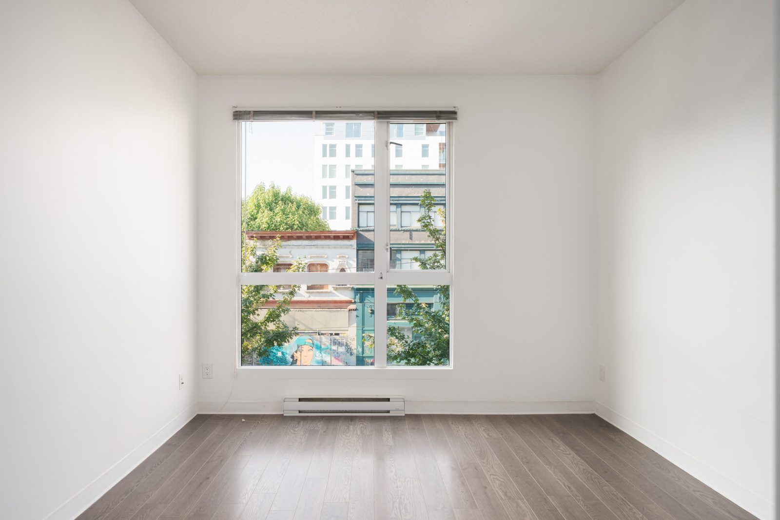 Empty room with white walls, dark wood floor, and large window showing trees and city buildings in Vancouver at 138 E Hastings St.