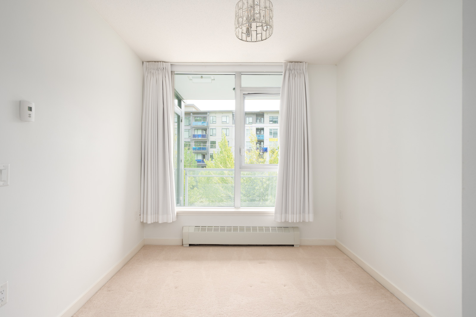 White room with beige carpet, ceiling light, and large window with white curtains facing apartments and trees on East Kent Ave Vancouver.
