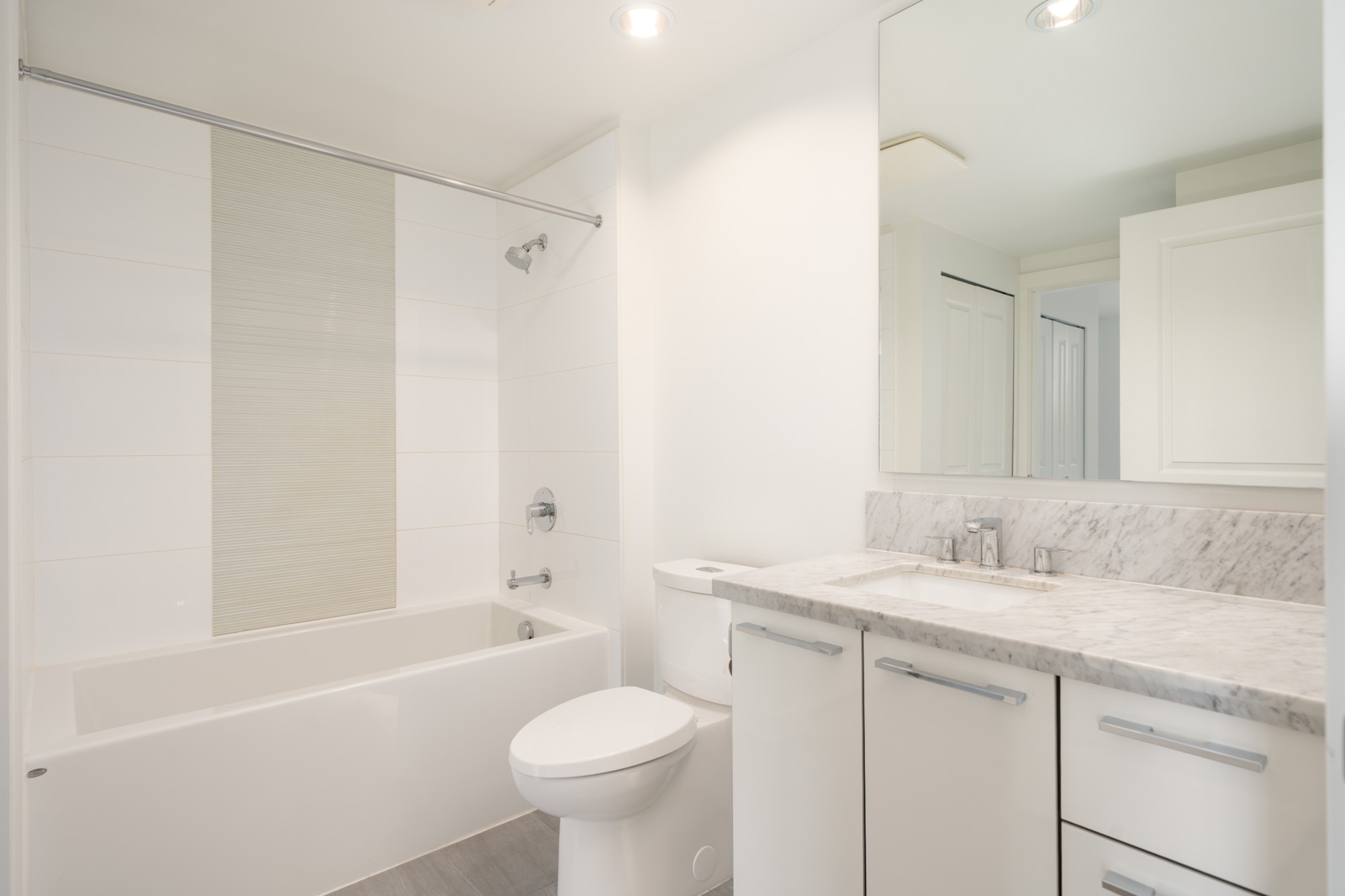 Modern bathroom with white walls, a bathtub-shower combo, toilet, large mirror, and marble-top vanity on East Kent Ave Vancouver.