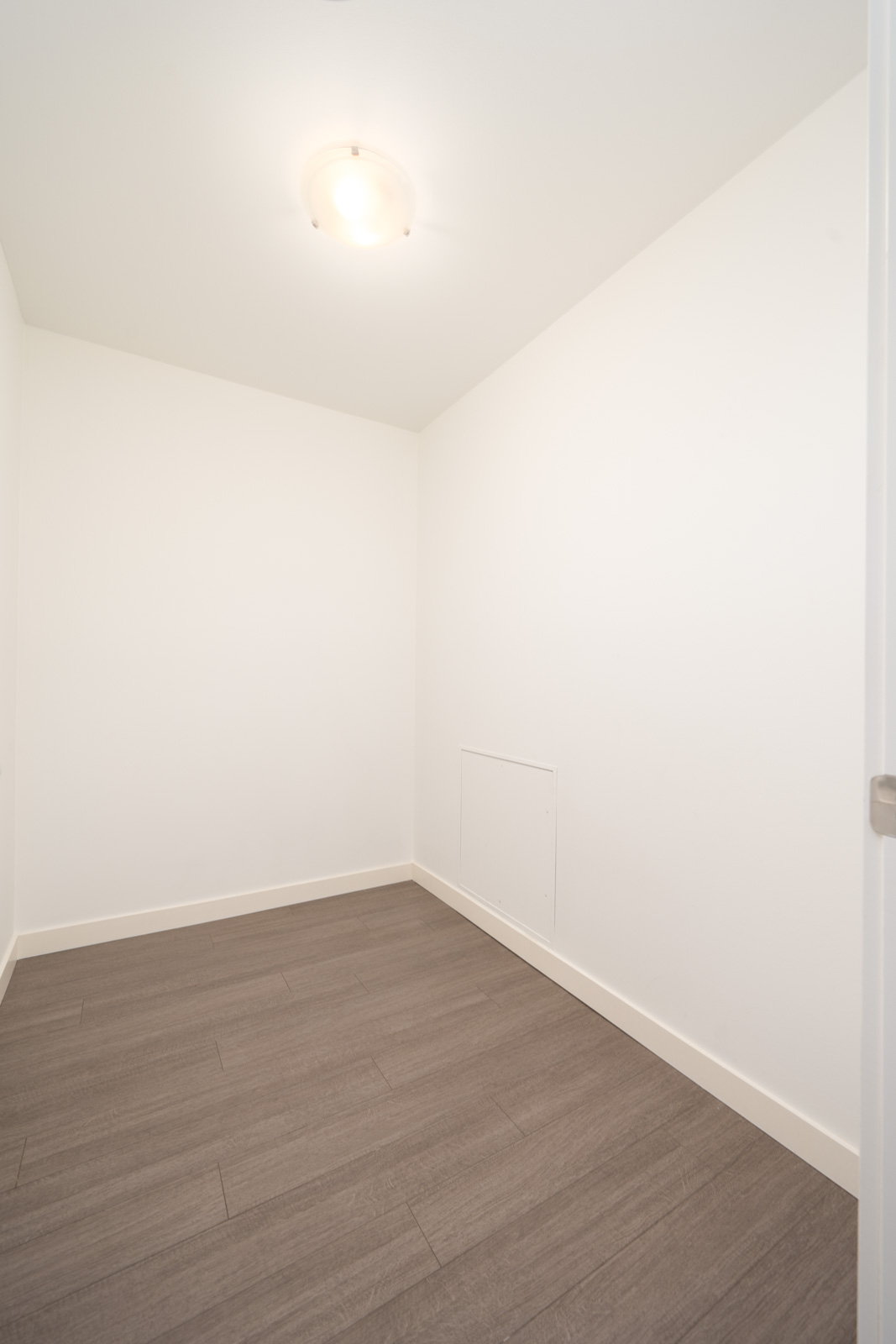 Empty room with white walls, ceiling light, gray wood floors, and a small wall access panel at 3281 East Kent Ave, Vancouver.