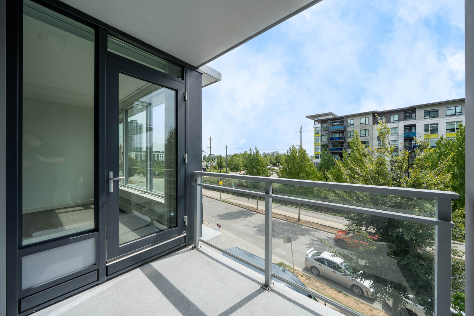 Balcony with glass railing overlooking East Kent Ave, Vancouver; street, parked cars, trees, and apartments under partly cloudy sky.