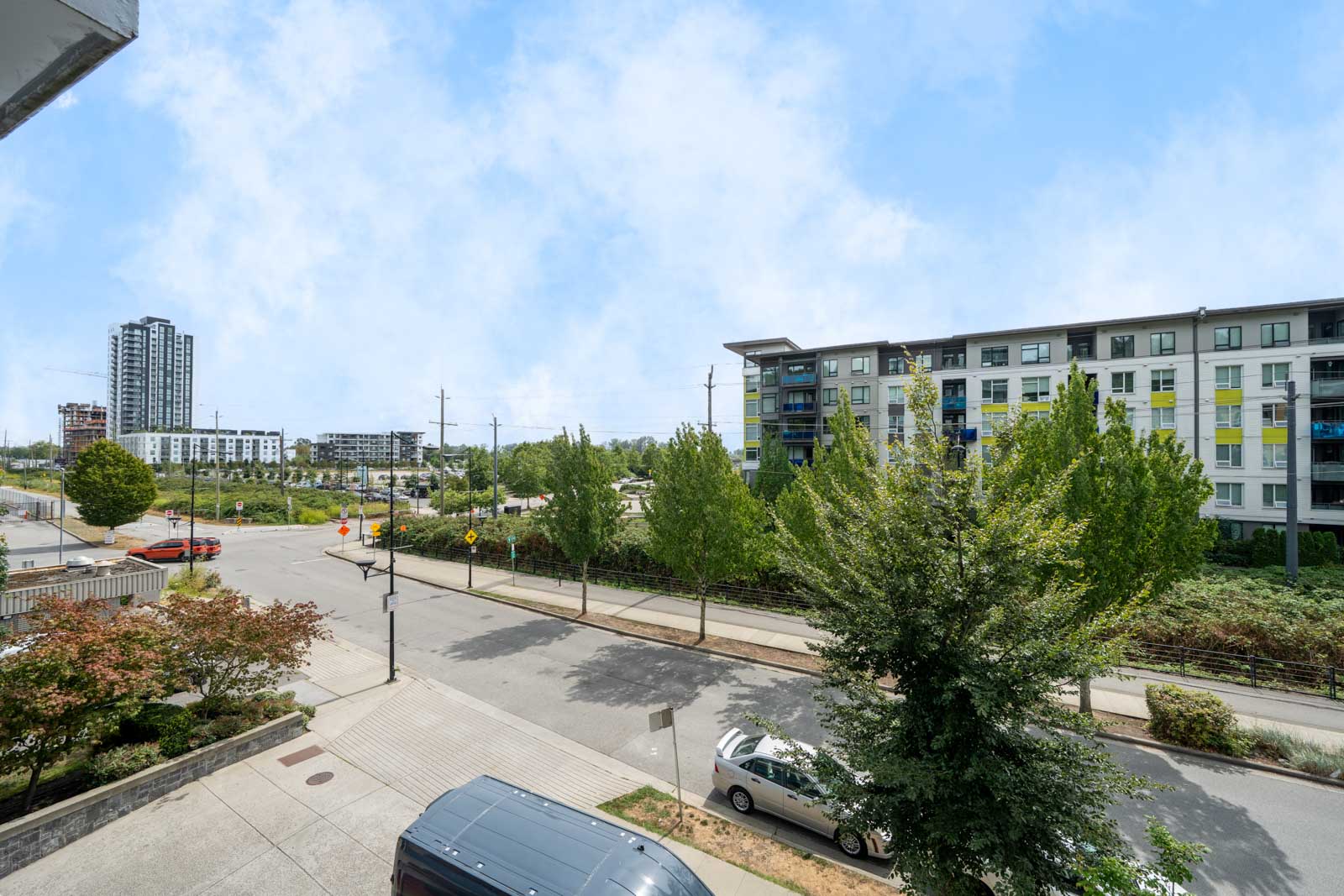 Quiet urban street on East Kent Ave, Vancouver, lined with parked cars, trees, and modern apartments beneath a lightly clouded blue sky.