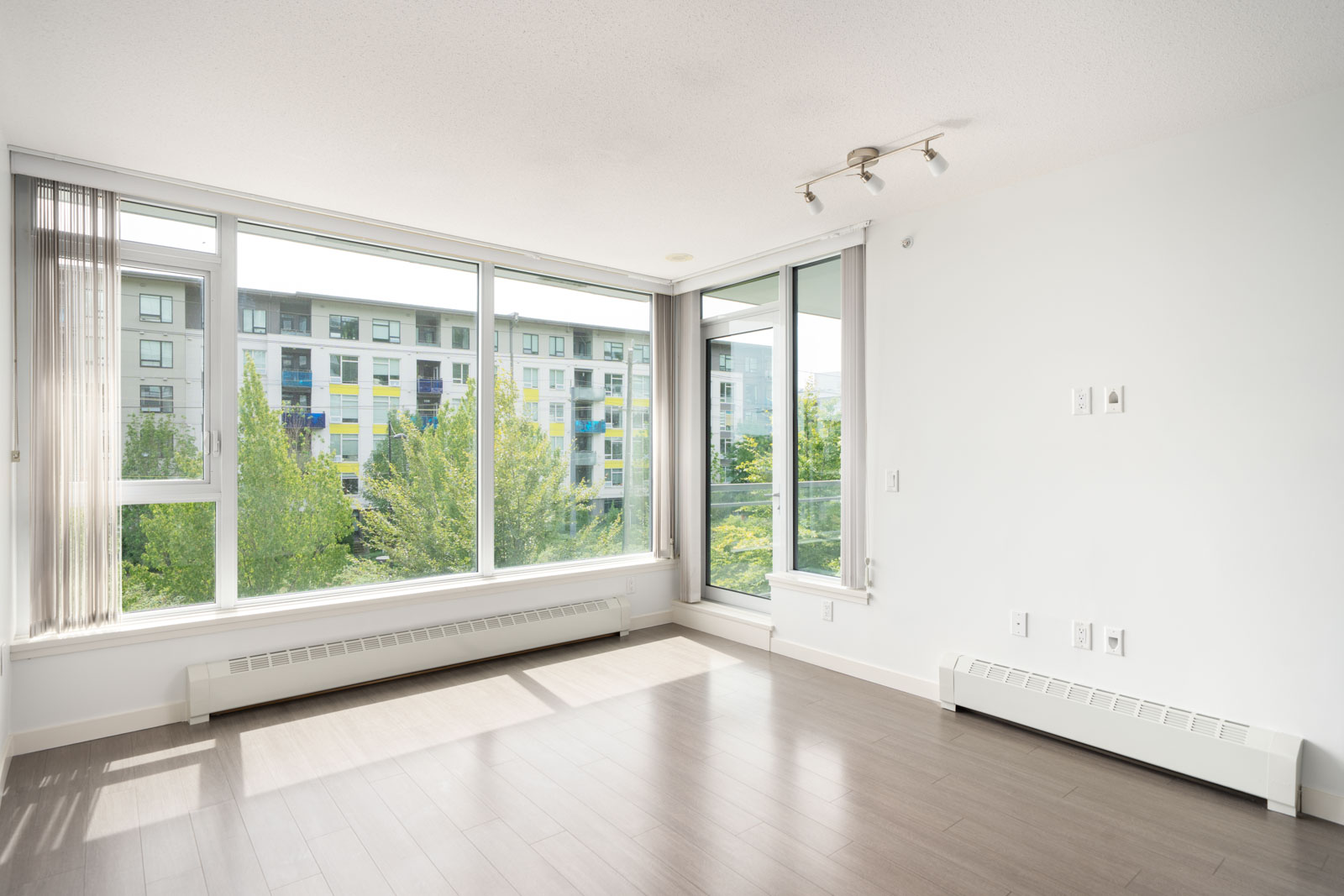 Bright, empty Vancouver apartment with large windows, light wood floors, white walls, and views of trees and a modern building.