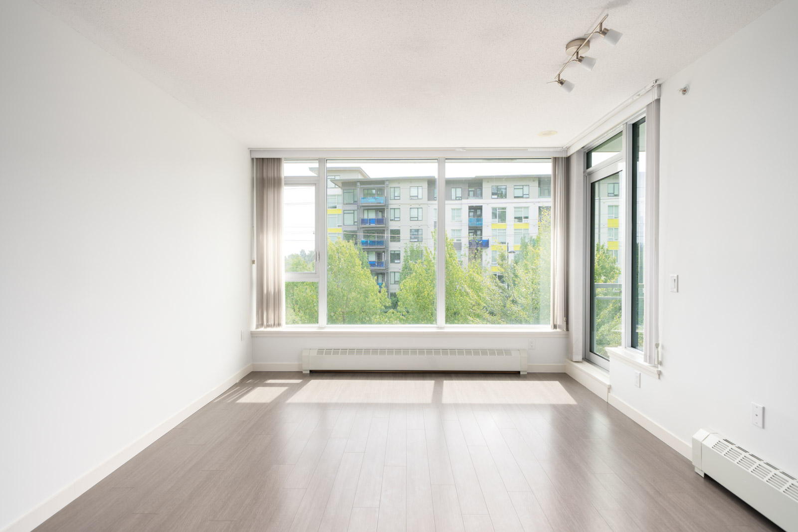 Empty room with large windows, white walls, light wood floors on 2nd floor at 3281 East Kent Ave, Vancouver; view of trees and apartments.
