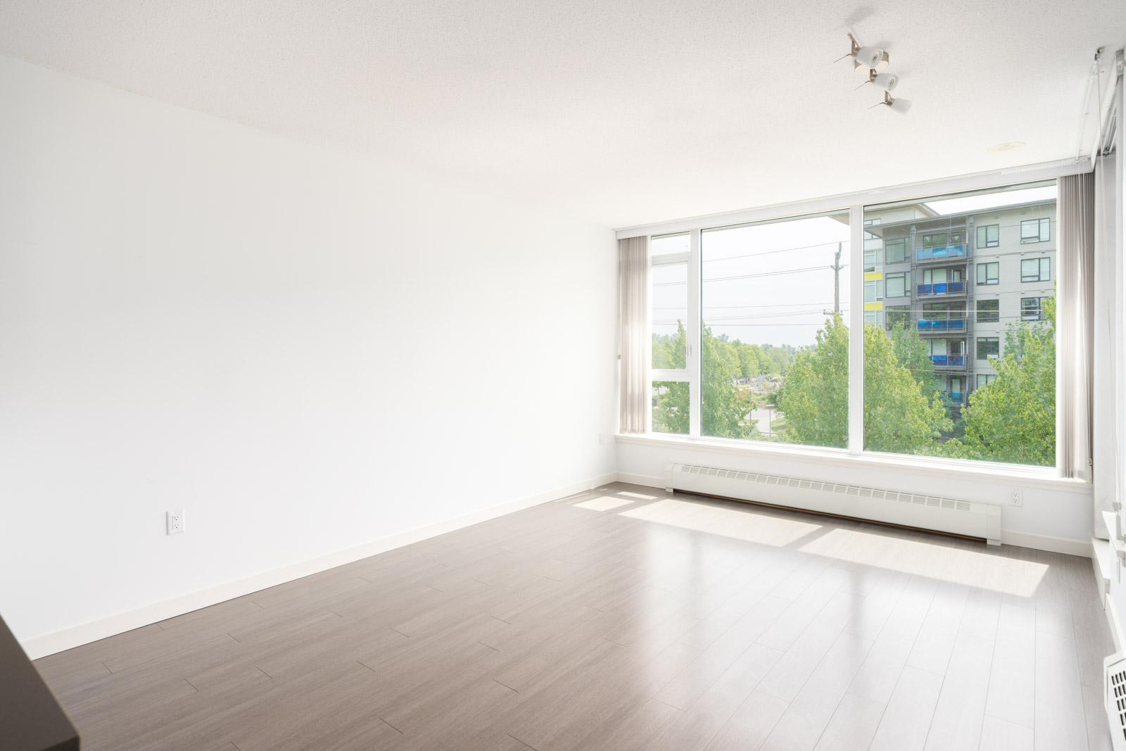 Bright, empty room with light walls, wood floors, and a large window facing trees and an apartment on East Kent Ave, Vancouver.