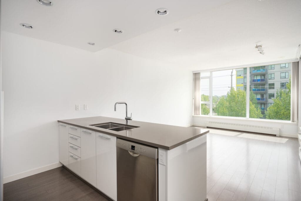 Modern, unfurnished kitchen with white cabinets, large island, stainless sink and dishwasher, open to living area with city views. Vancouver.