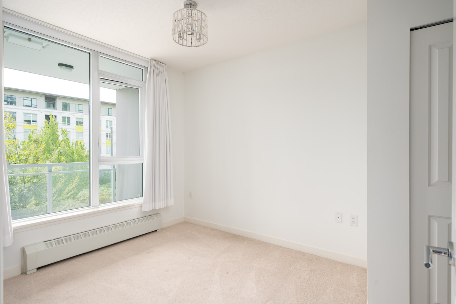 Empty, carpeted room with white walls, a large curtained window showing trees and an apartment building. Located in Vancouver.