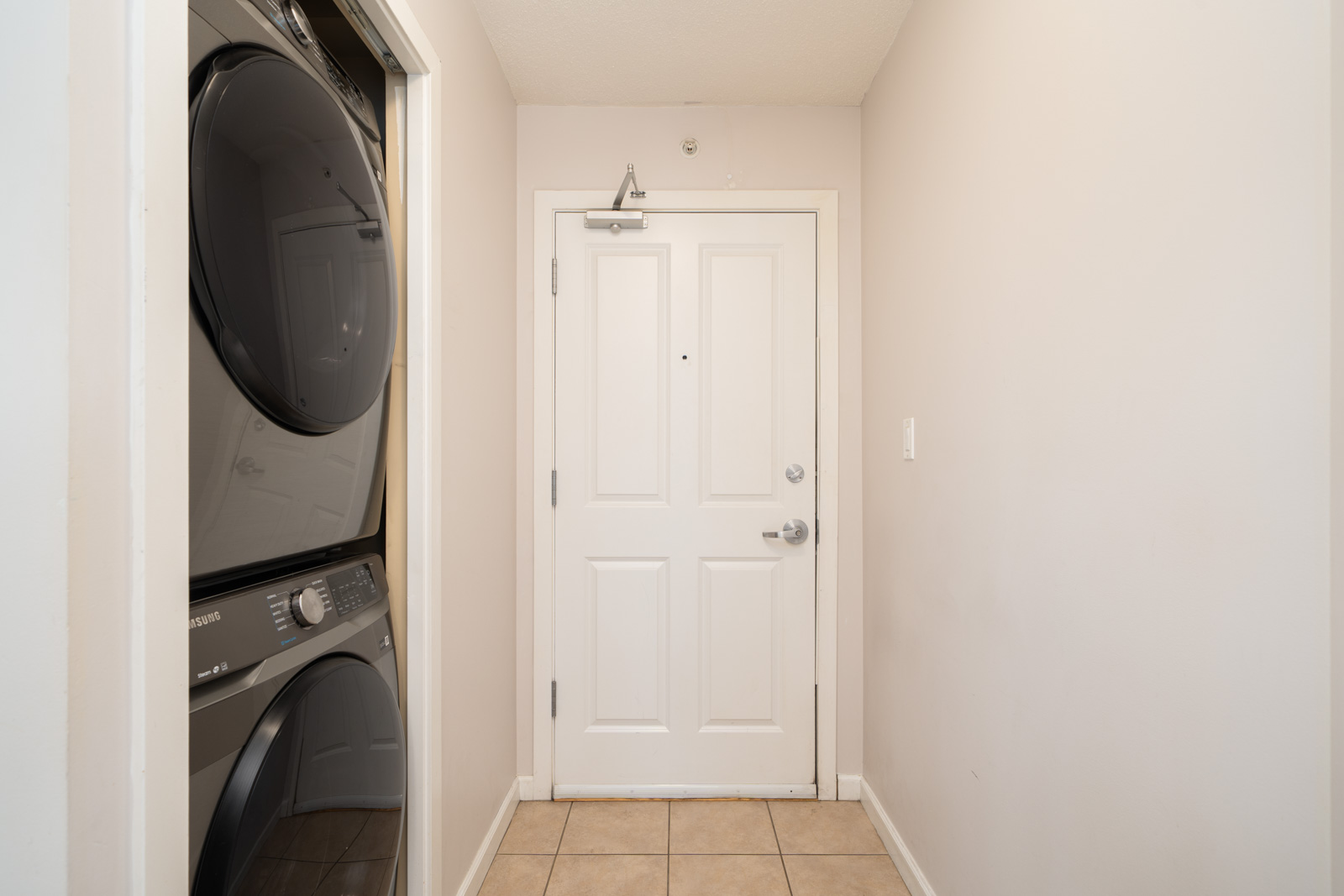 A white front door with a silver handle sits at the end of a tiled hallway; stacked black washer and dryer in an alcove left.