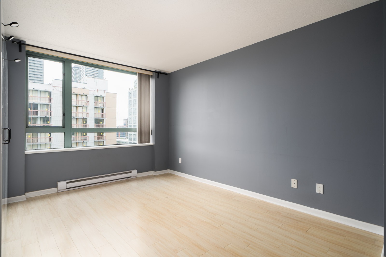Unfurnished Burnaby condo room on Halifax St with light wood floors, blue-gray walls, city-view window, and baseboard heater.