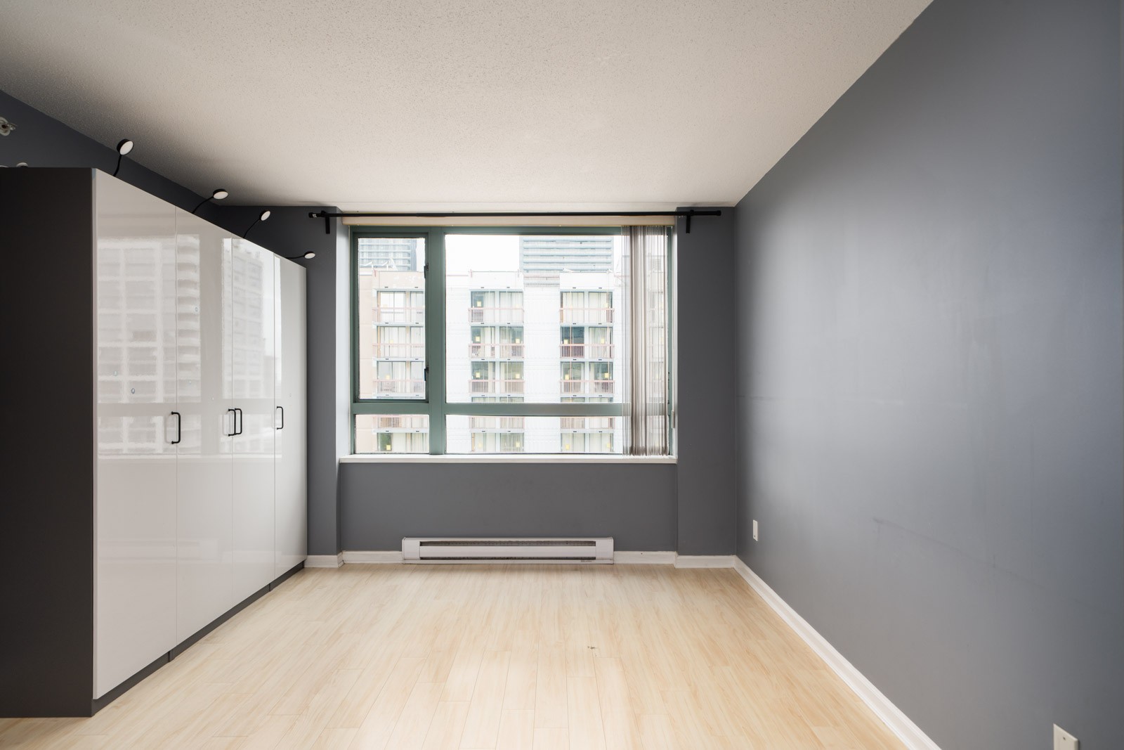 Empty room with light wood floors, dark gray walls, a large window with a city view, and white built-in cabinets on the left at Halifax St Burnaby.