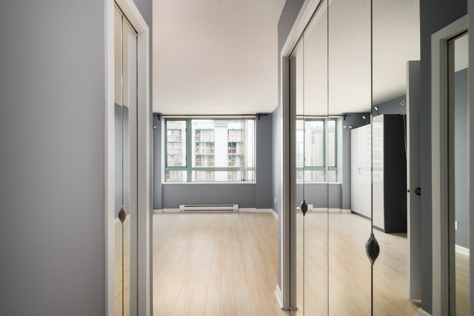 Sunny, empty room with light wood floors, large windows, and mirrored closet doors in a 16th floor Halifax St Burnaby condo.
