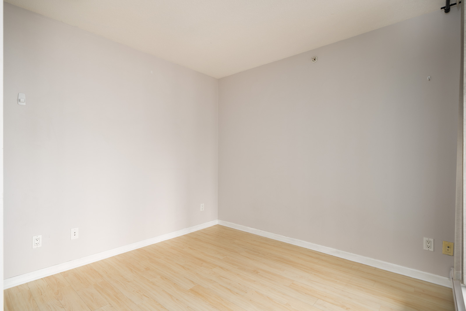 Unfurnished corner of a Burnaby condo room with light beige walls, light wood floor, and outlets along the baseboards.