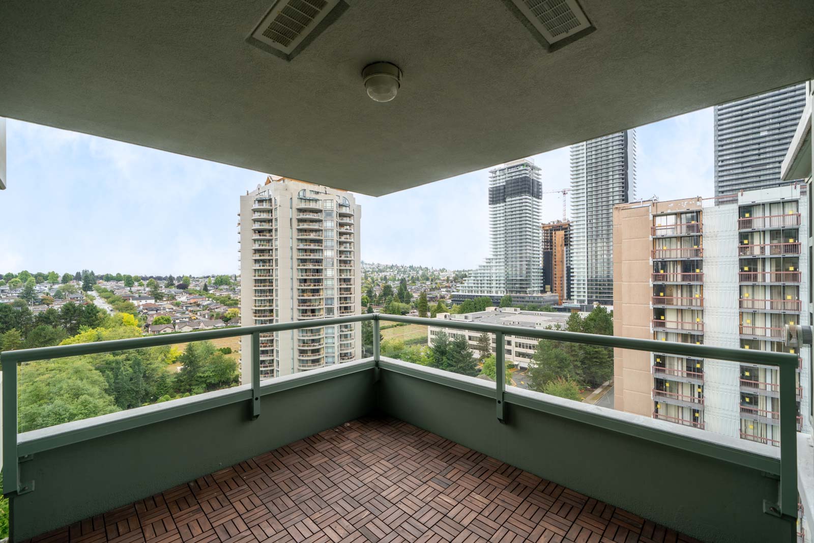 Balcony with tile floor and glass railing on 16th floor, overlooking Burnaby city buildings, apartments, and greenery under clouds.