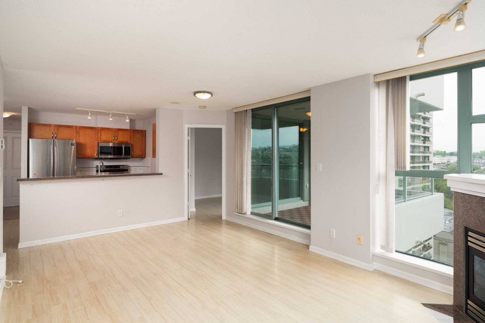 Unfurnished 16th floor condo living area in Burnaby with light wood floors, open kitchen, balcony access, windows, and fireplace.
