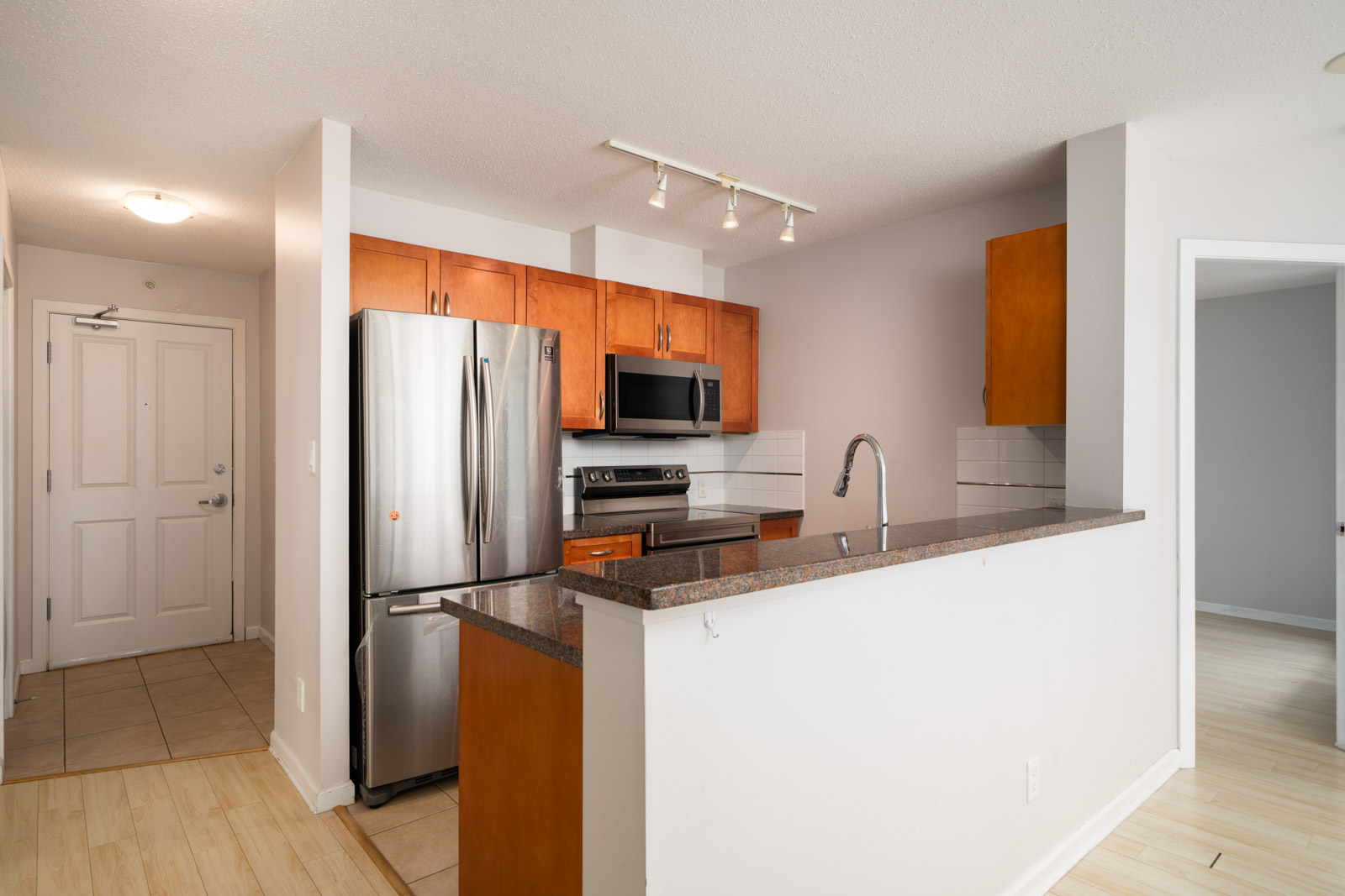 Modern kitchen with stainless steel appliances, wood cabinets, granite counters, tile floor, and nearby hall in a 16th-floor Burnaby condo.
