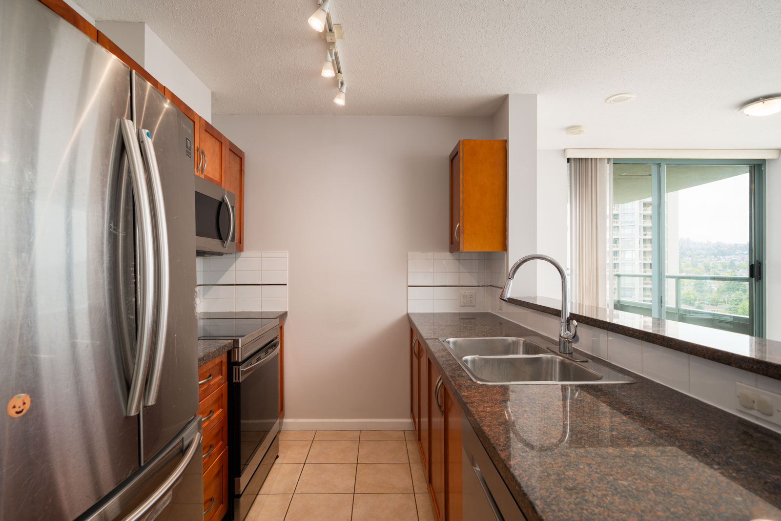 Modern kitchen in a Burnaby condo with stainless appliances, brown cabinets, double sink, tile floor, and balcony window.