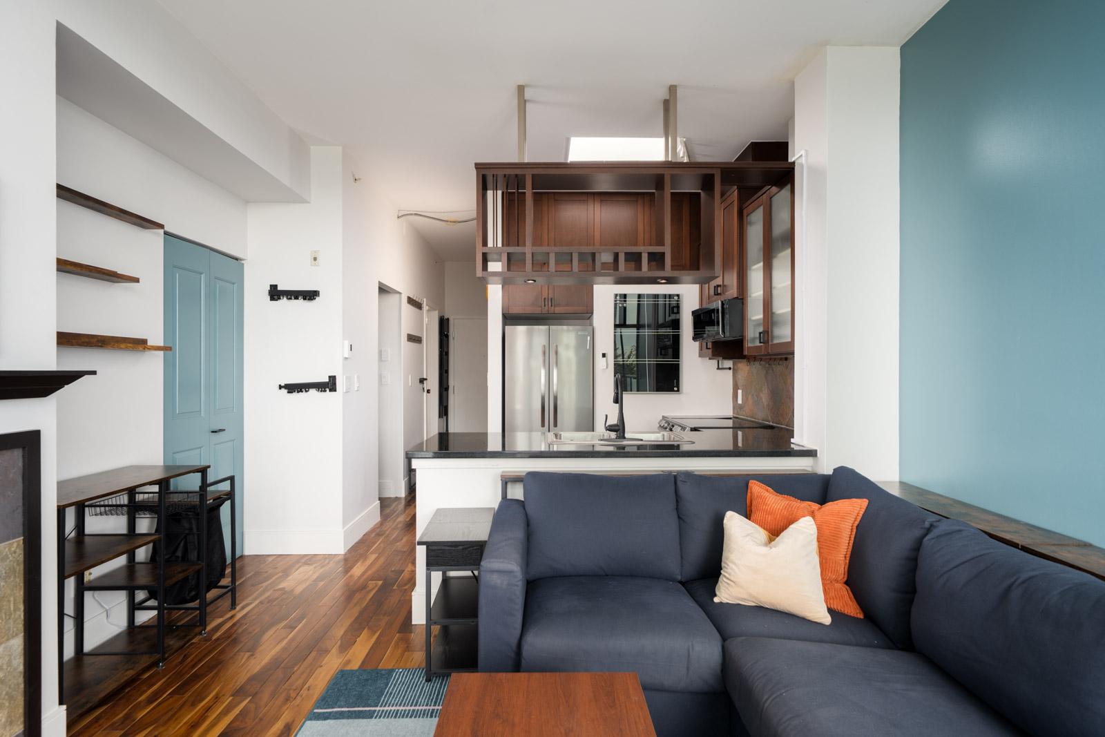 Modern open-concept living area on Hudson St, Vancouver with blue sofa, orange and cream pillows, hardwood floors, and stainless kitchen.