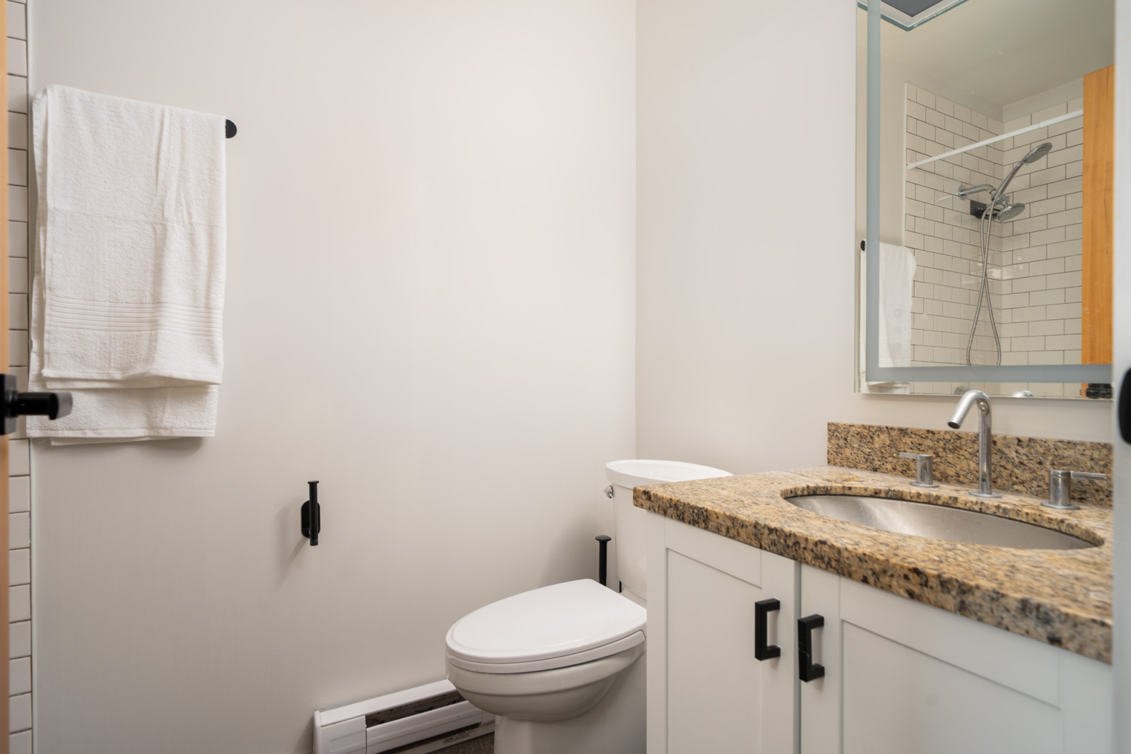 Modern bathroom on Hudson St, Vancouver with granite countertop, white cabinets, towel rack, toilet, and shower seen in mirror.