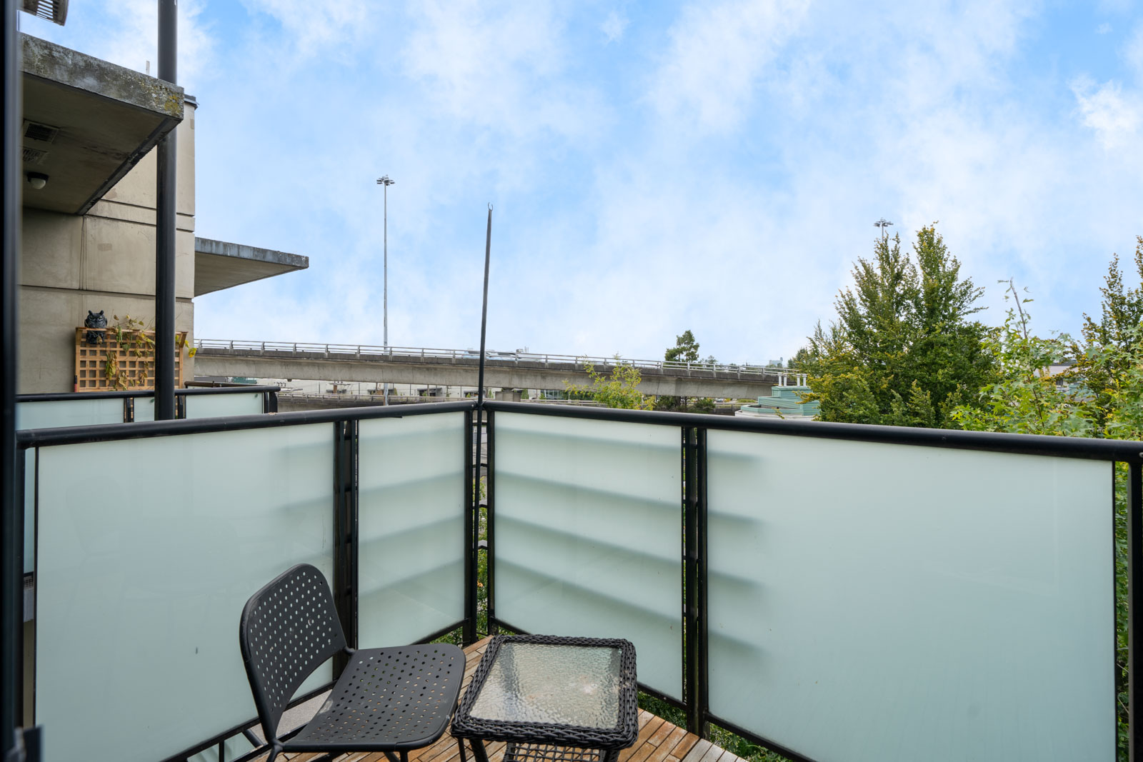 Small balcony at 8988 Hudson St, Vancouver, with frosted glass railing, two black chairs, table, and view of highway and trees.