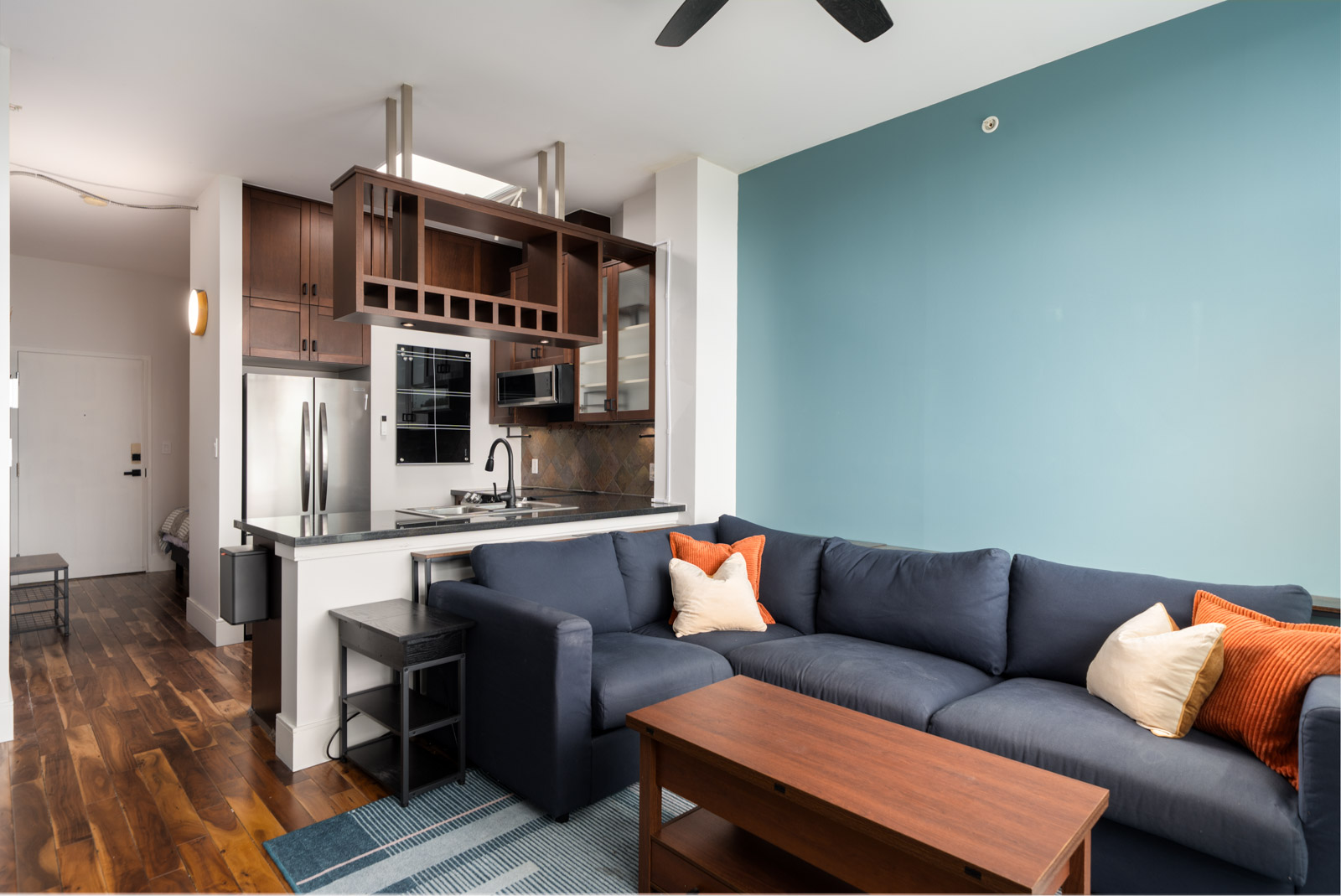 Open-plan living room and kitchen on Hudson St, Vancouver, with a blue accent wall, dark blue sofa, wood table, and steel appliances.