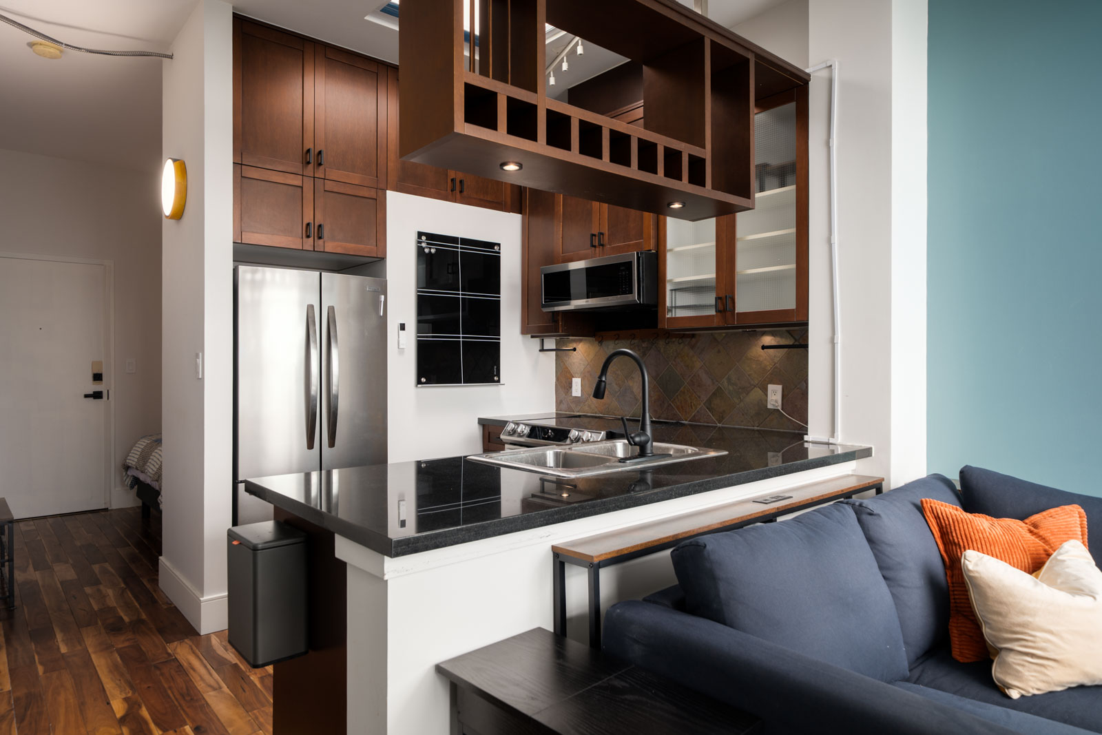 Modern kitchen with stainless appliances, dark wood cabinets, black countertop, blue sofa with orange pillow in Vancouver.