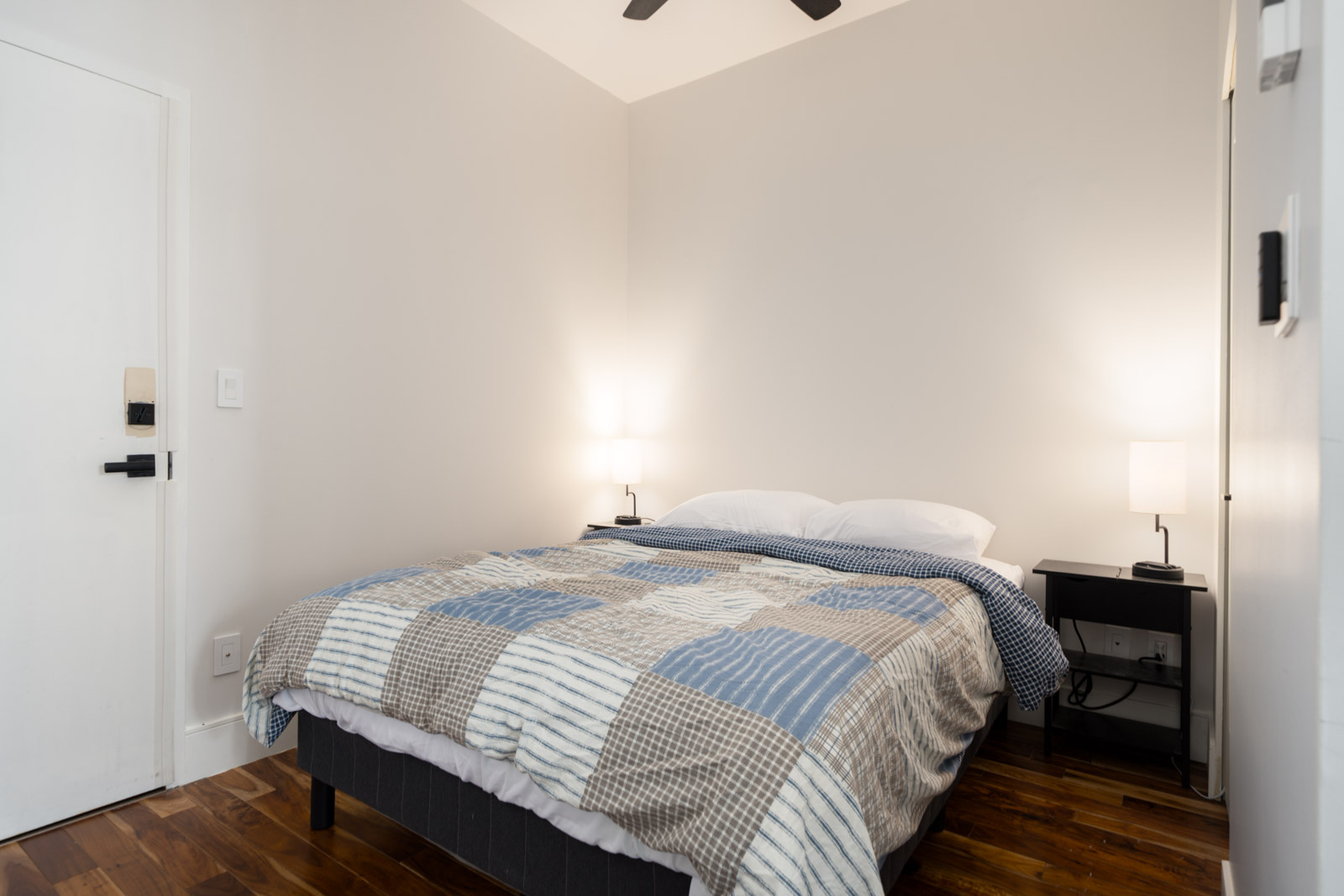 A neatly made bed with a patterned comforter stands between two black nightstands in a Hudson St, Vancouver bedroom with hardwood floors.