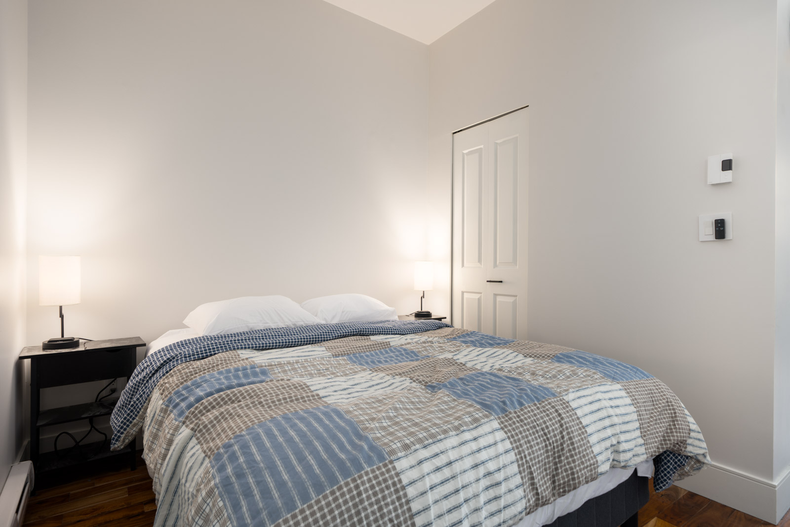 A minimalist Hudson St, Vancouver bedroom with a neatly made bed, patterned quilt, black nightstands, and lamps against a pale wall.