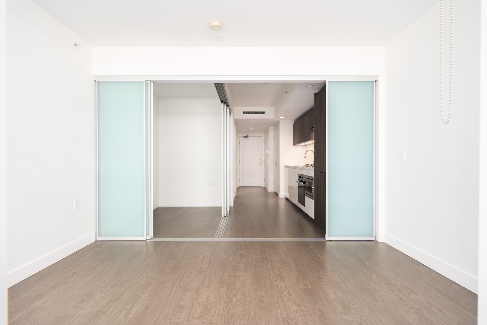 Modern empty apartment room on 6th floor in Richmond with light wood floors, white walls, glass doors, and kitchen with dark cabinets.
