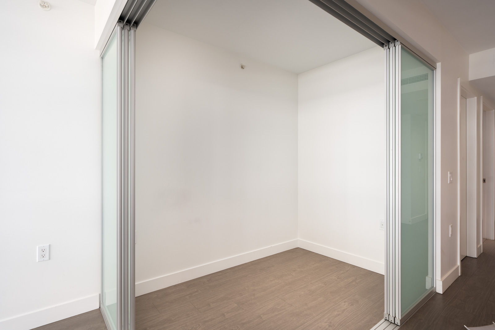 Small empty room with white walls, light wood flooring, and partly open sliding glass doors; hallway at right. 6th floor, No.3 Rd, Richmond.