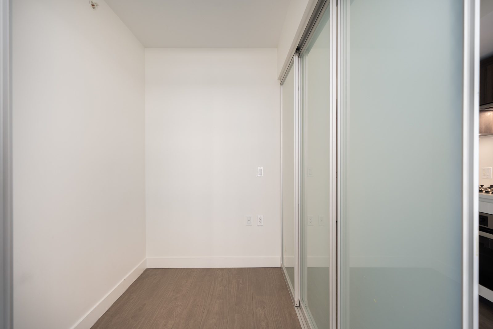 Small empty room with white walls, light wood floors, sliding glass doors right side, next to a kitchen; Richmond, 6320 No.3 Rd.