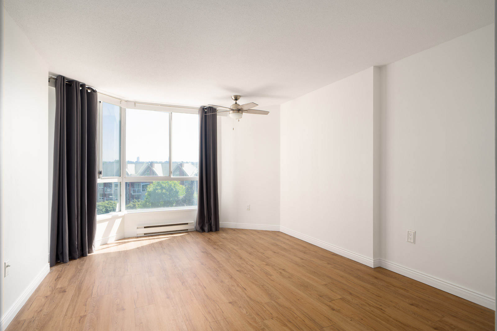 Empty room with wood floors, white walls, large window with black curtains, ceiling fan, and baseboard heater. Sunlight fills the space on Quayside Dr, New Westminster.